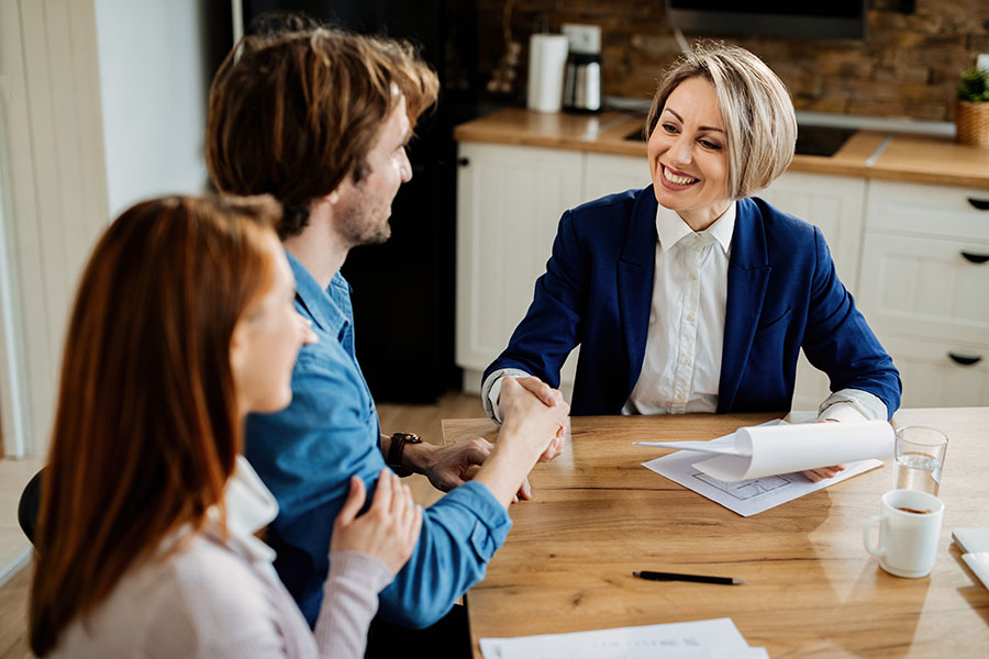 About Our Agency - Portrait of a Cheerful Mature Insurance Agent Holding Paperwork and Shaking Hands With a Young Couple As They Sit in Their New Home